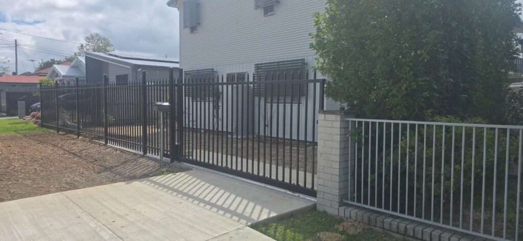 Black Zeus spear top aluminium sliding gate closed across a concrete driveway, in line with the matching spear top fence panels beyond, custom welded by Accolade Fencing